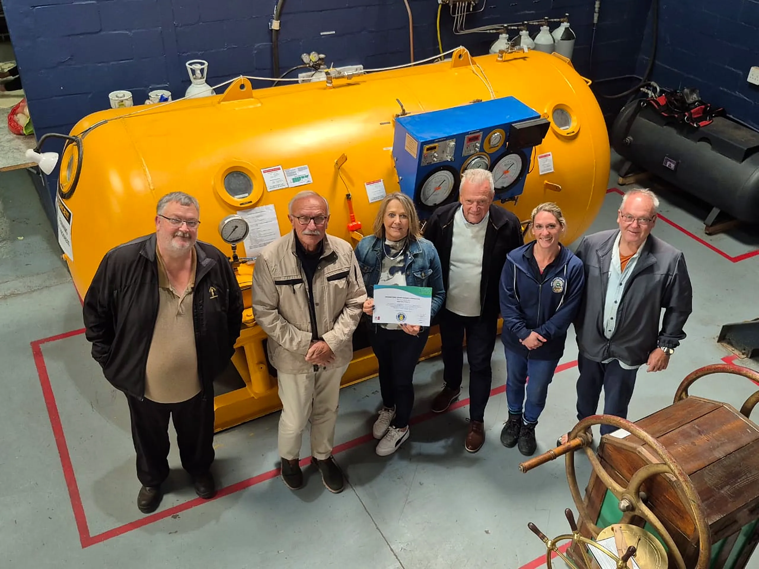 jacks-dive-chest-idsa-full-member-team Jacks Dive Chest team and visitors beside a diving chamber holding the IDSA certificate
